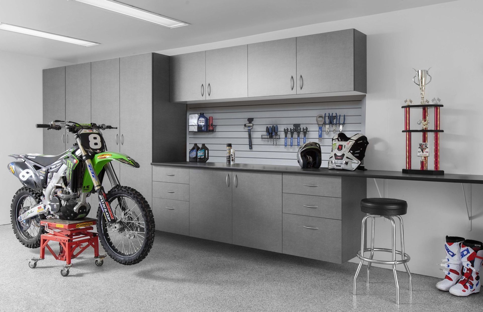 A motocross bike on a stand next to gray garage cabinets, a workbench with tools, a trophy, and boots on a speckled floor.