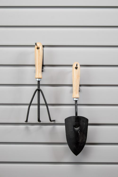 Two small gardening hand tools, a cultivator and a trowel, hang on a grey horizontal slat wall.