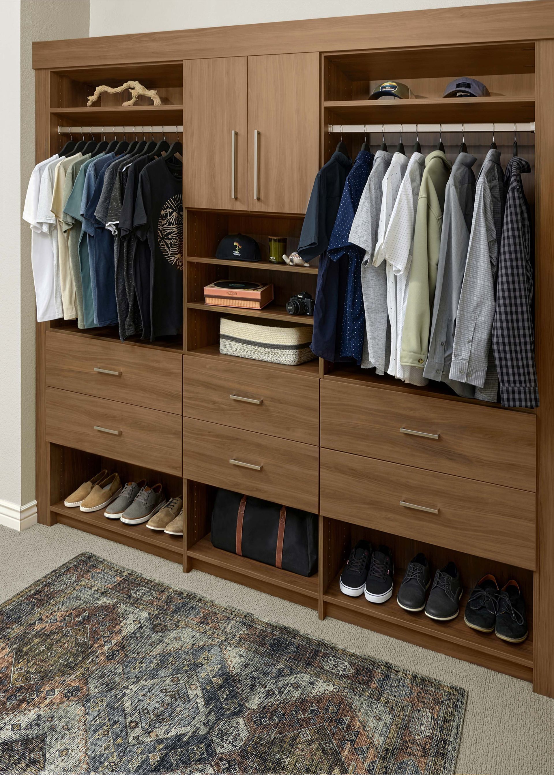 A wood-finished closet organizer with hanging rods, drawers, open shelving, and a patterned rug on the floor.