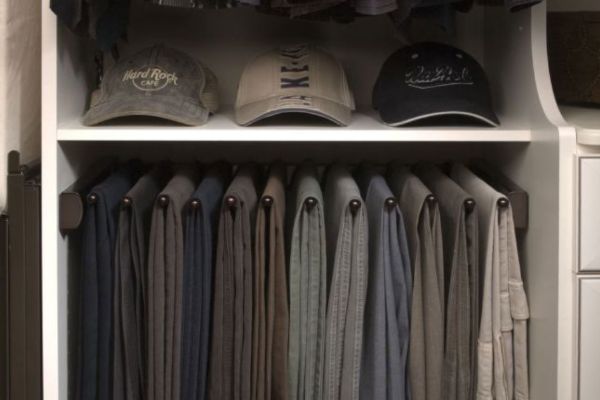 A closet shelf featuring three baseball caps above a rack organized with several pairs of folded hanging pants.