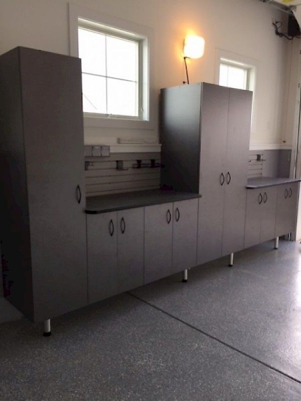 A garage storage system with dark gray cabinets, a black countertop, and mounted wall slats beneath two windows.