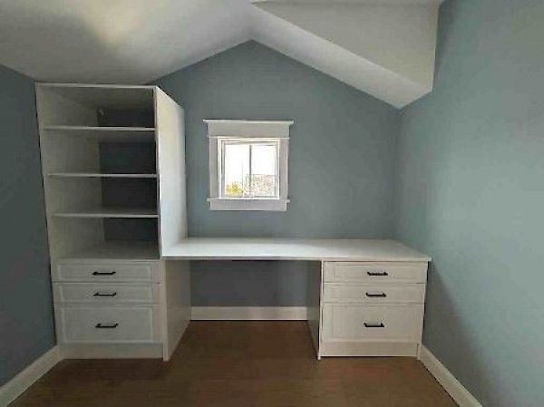 A white built-in desk and shelving unit with drawers sits against a blue wall under a sloped ceiling with a small window.