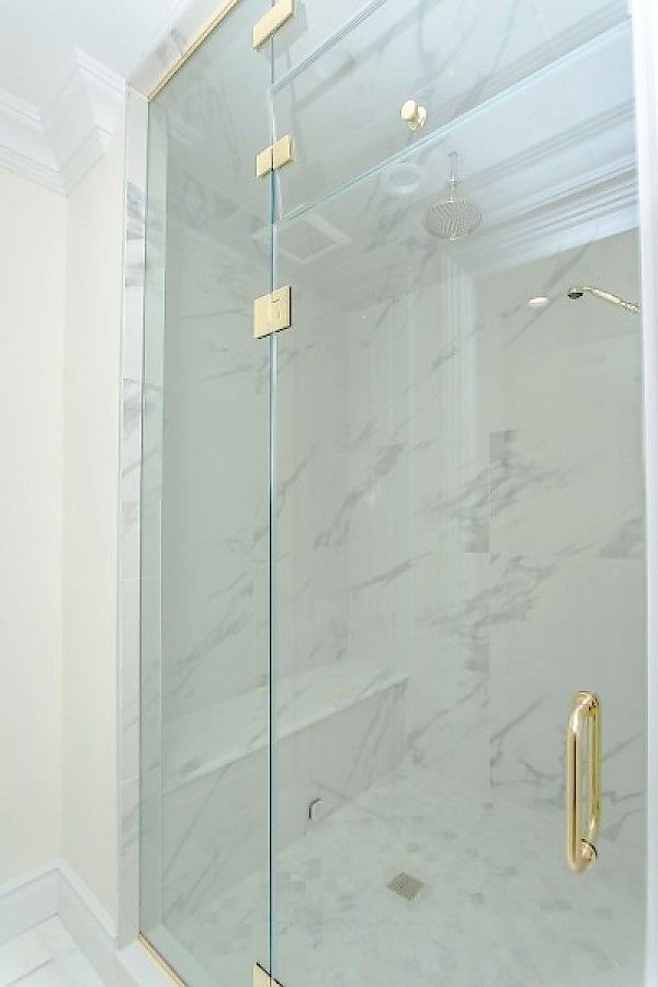 A glass shower door with gold hinges and a gold handle, set against white marble walls inside a bathroom.