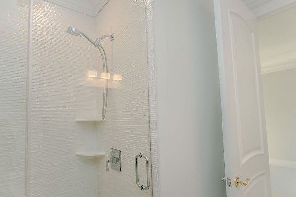 A modern bathroom shower featuring textured white walls, a silver showerhead, built-in corner shelves, and a door nearby.
