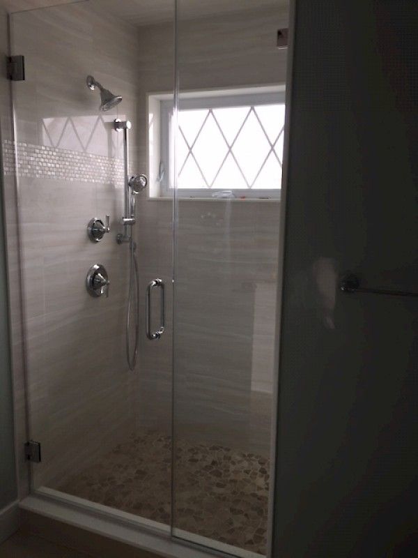 A glass-enclosed shower with tiled walls, a handheld showerhead, a pebble floor, and a small diamond-patterned window.