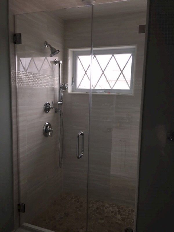 A modern walk-in shower with light tile walls, a glass door, a hand-held shower head, and a small diamond-pattern window.