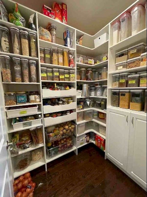 A well-organized white pantry with shelves filled with clear storage containers, wire baskets of produce, and a bag of onions.