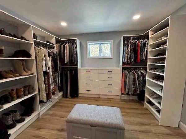 Walk-in closet with white shelving units, hanging clothes, drawers, and a gray ottoman on wood-look flooring.