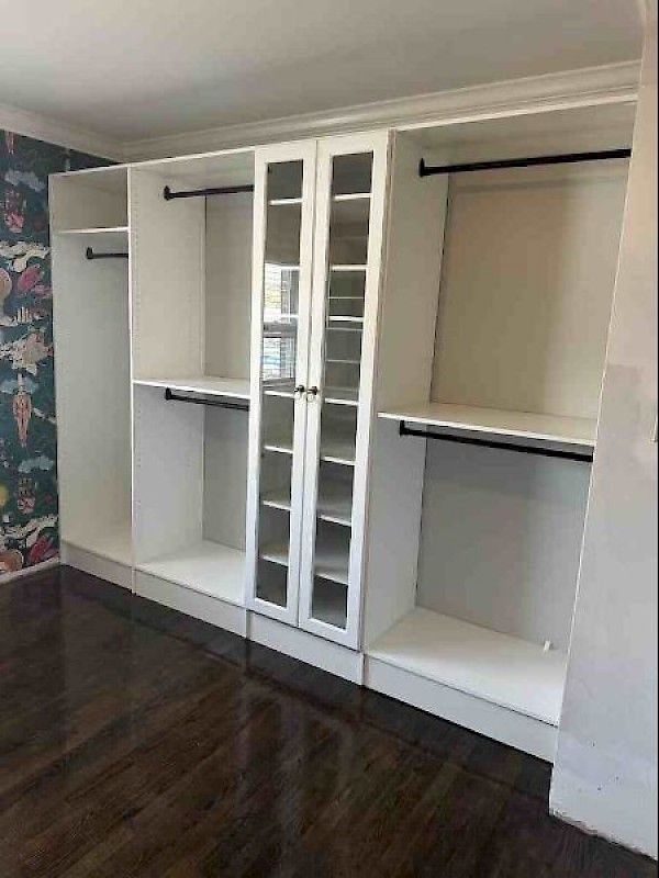 A large, white, built-in closet unit with open hanging sections and a central glass-paned cabinet on a dark wood floor.