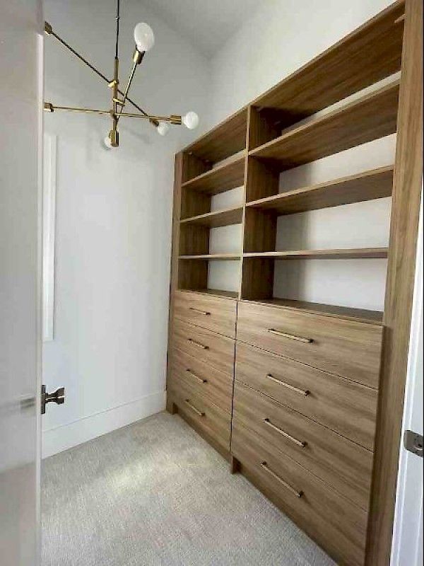 A walk-in closet featuring light wood built-in shelving, drawers with gold handles, beige carpet, and a gold chandelier.