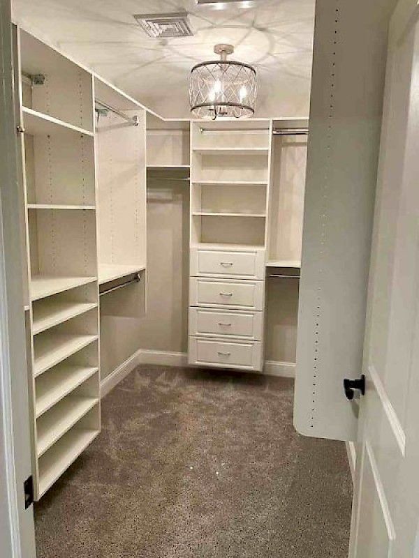 A walk-in closet featuring white wooden shelving, drawers, and hanging rods against beige walls with gray carpet flooring.