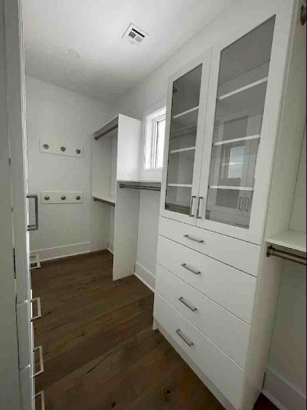 A white walk-in closet featuring floor-to-ceiling cabinetry with glass doors, drawers, hanging rods, and dark wood floors.