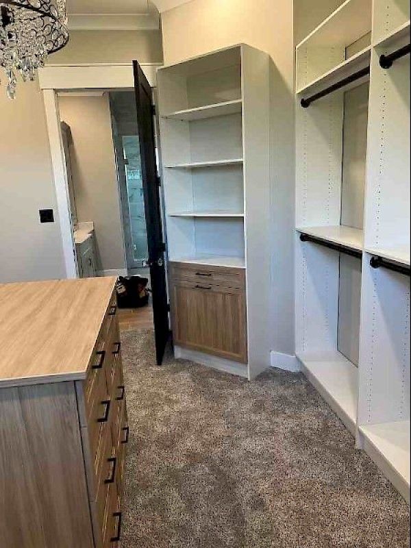 A walk-in closet featuring a wood-toned dresser, white shelving units with a lower cabinet, and grey carpeting.
