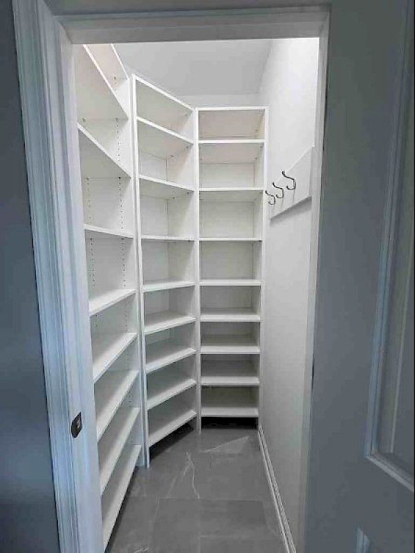 A small, narrow walk-in pantry with white built-in shelving units lining three walls and a row of hooks on the right side.