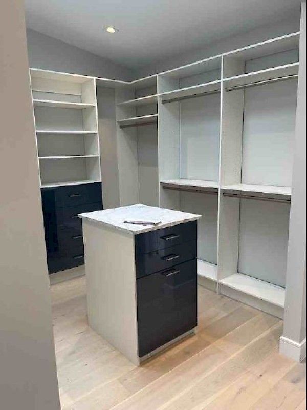 A walk-in closet featuring white shelves and hanging rods, with a central island and drawers in glossy dark navy finish.