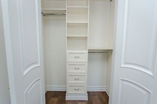 An open closet featuring white shelves and four drawers, with hanging rods on both sides and a dark wood floor.
