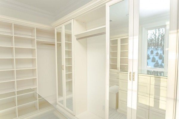 A walk-in closet with white shelving, a hanging rod, glass-front cabinets, and a mirror reflecting a window.