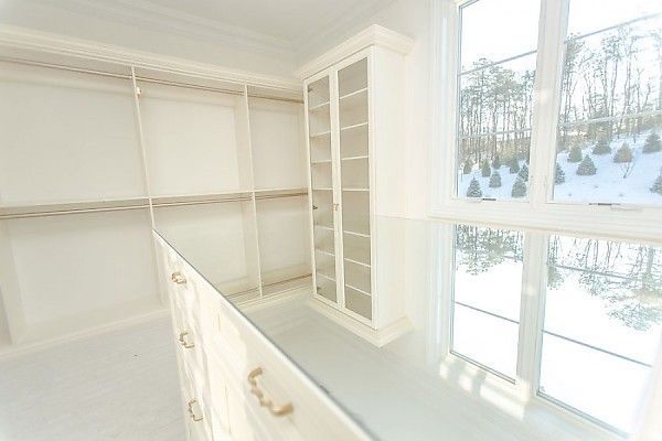 A bright, white walk-in closet featuring built-in shelving, a glass-topped center island, and a window with a snowy view.