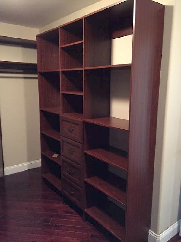 A tall, dark cherry wood modular closet shelving unit featuring open shelves and a central column of four drawers.