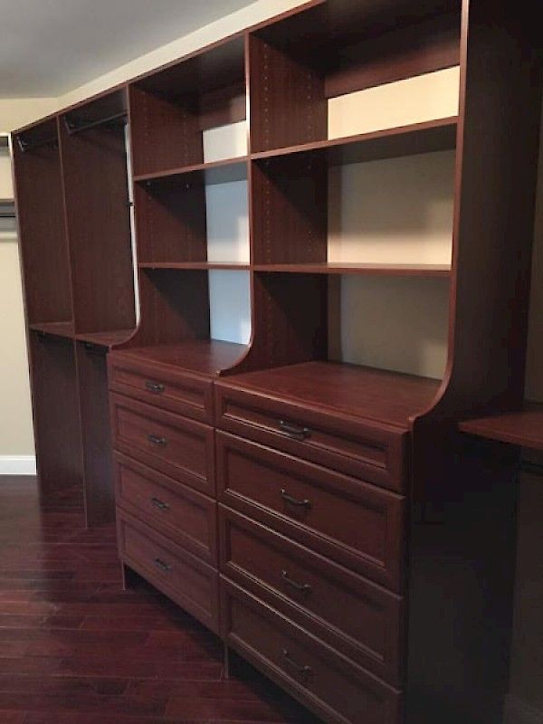 Custom dark wood walk-in closet shelving unit with built-in drawers and hanging rods on a dark hardwood floor.