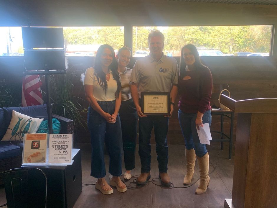 Group of five people holding a certificate, smiling, indoors with blue sofas, and a corrugated metal ceiling.