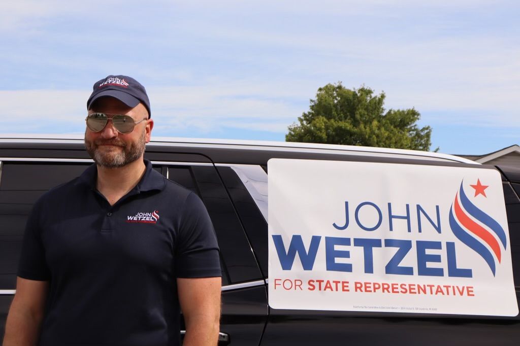 A man is standing in front of a van with a sign that says john wetzel for state representative
