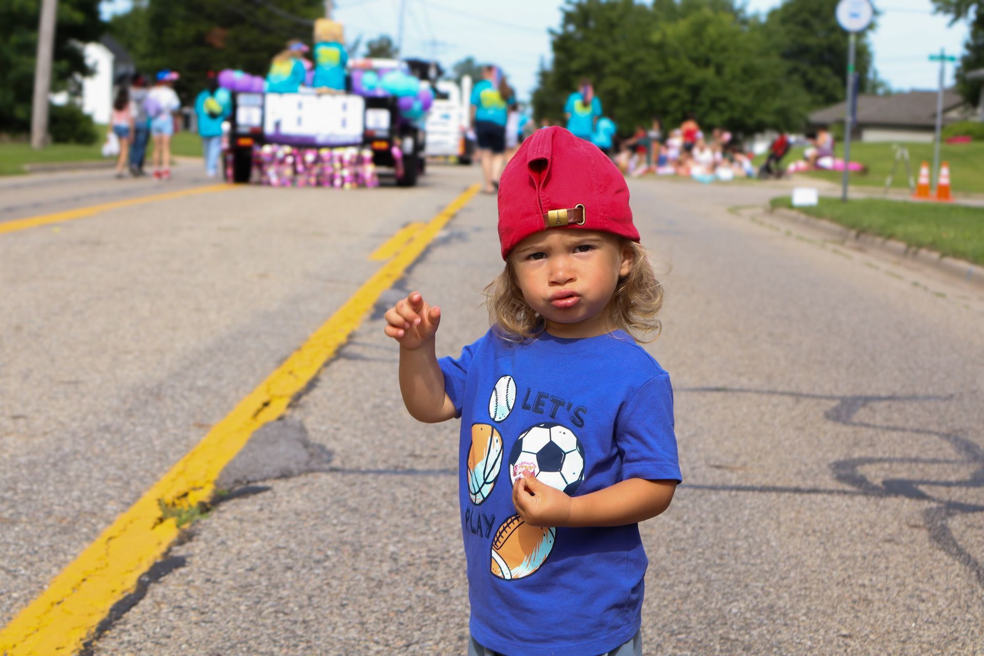 A little boy wearing a blue shirt and a red hat is standing on the side of the road.