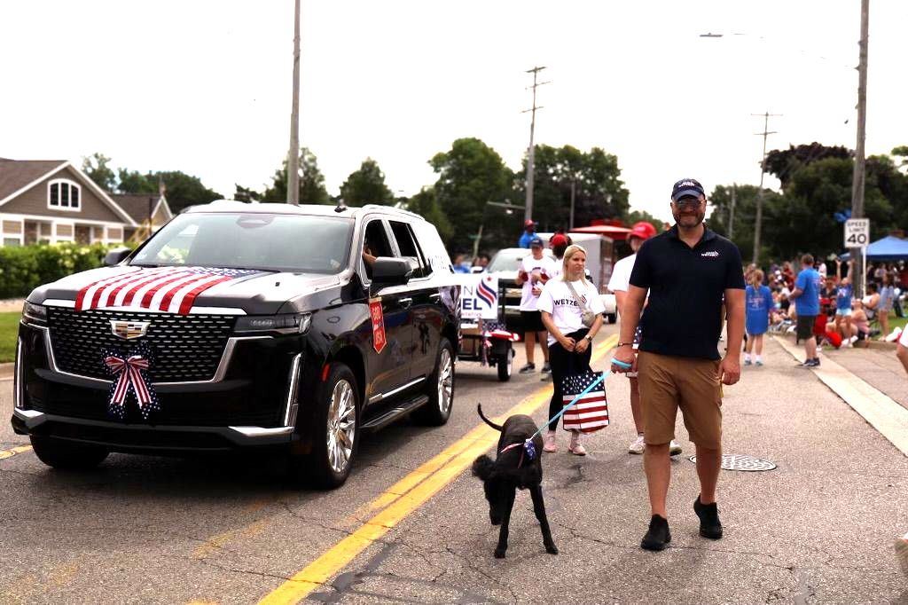 A man and a dog are walking down the street in front of a car in a parade.