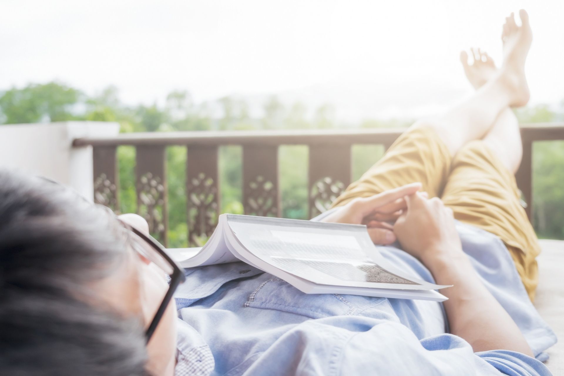 A person resting on a balcony with their feet up and an open book resting on their chest.