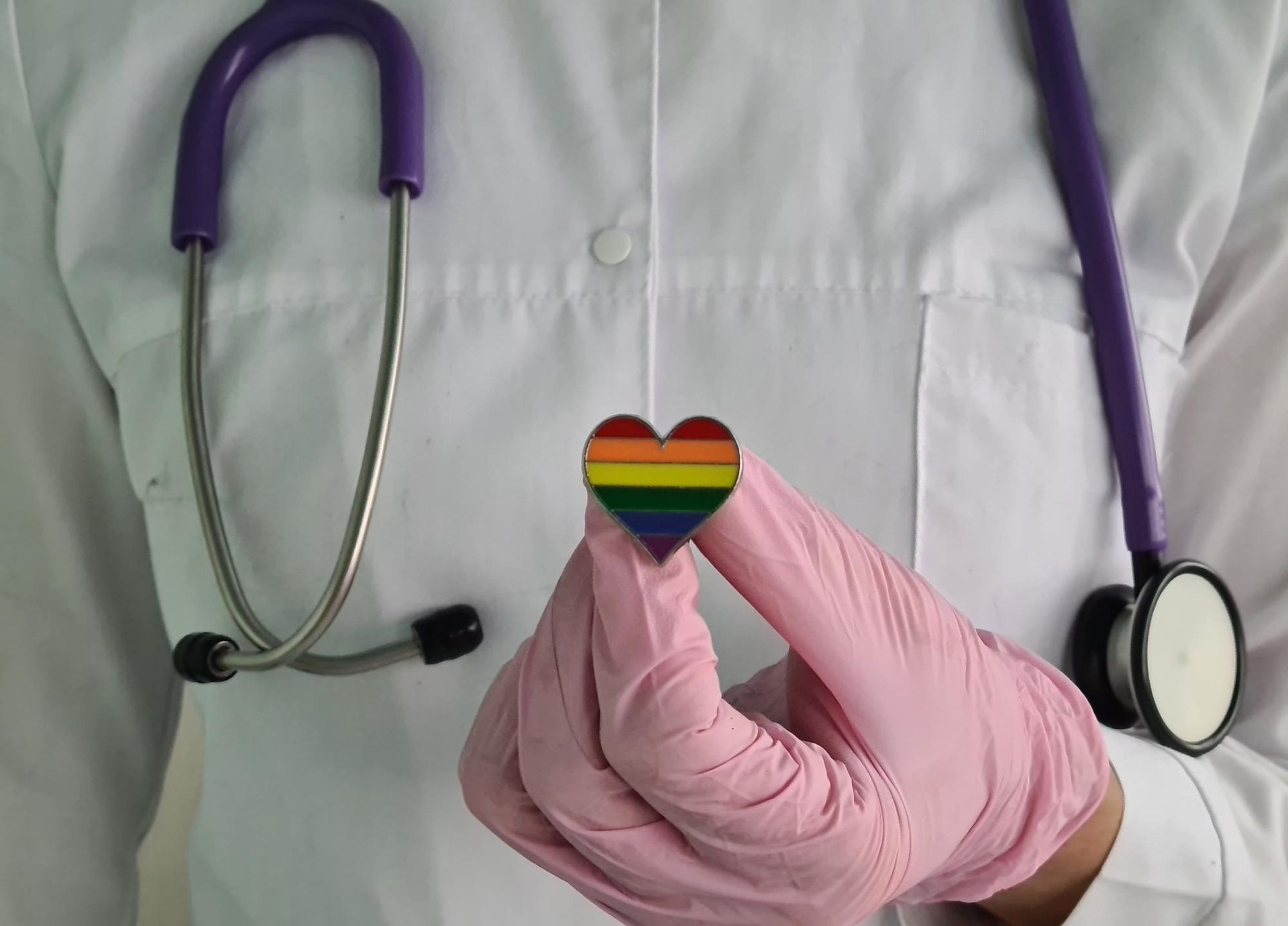 A person in a white lab coat and pink gloves holds a small rainbow heart pin, with a stethoscope draped around their neck.