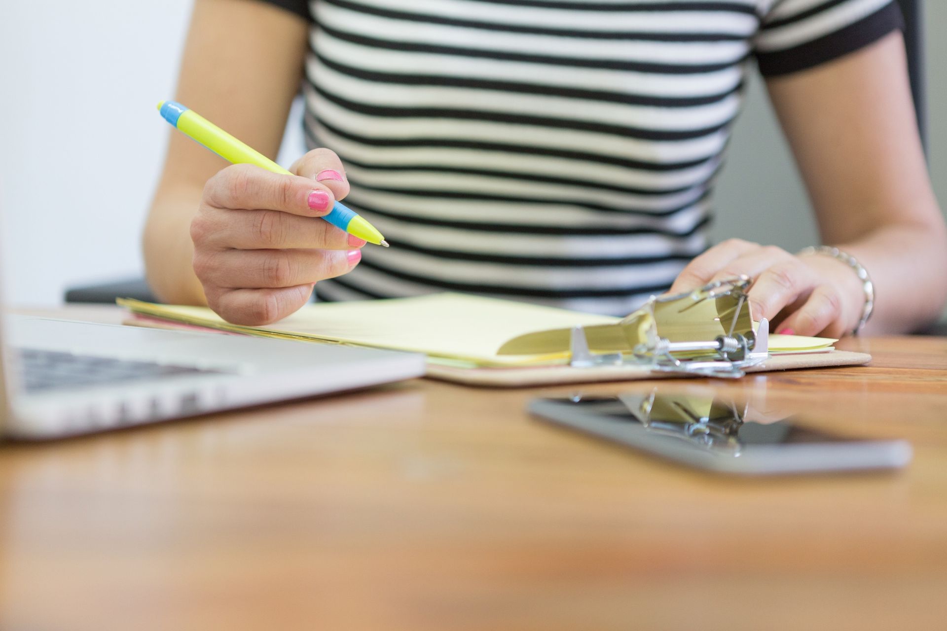 A person in a striped shirt writing on a clipboard with a yellow pen, with a laptop and smartphone nearby.