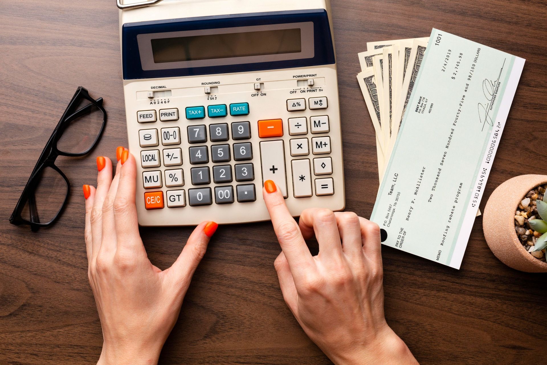 Hands resting on a wooden desk, using a calculator next to bank checks, cash, and glasses.