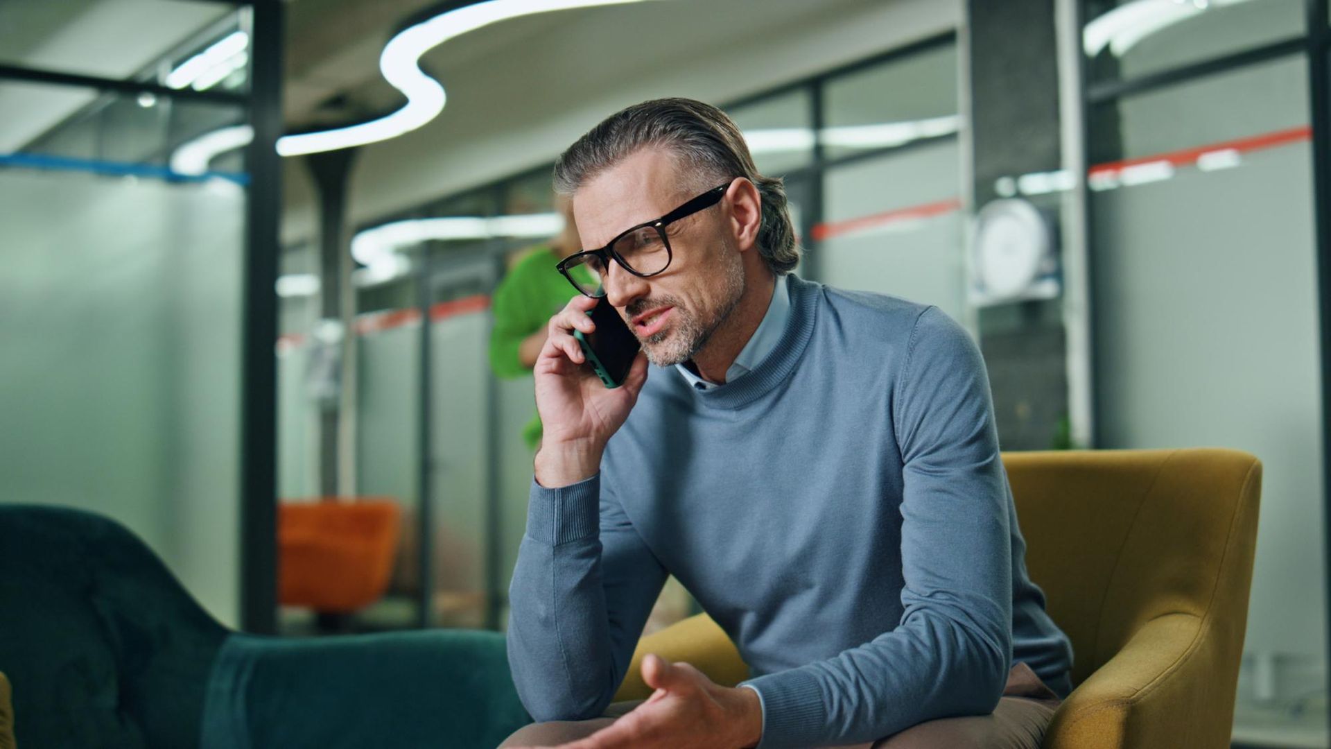 A man wearing a blue sweater and glasses talks on his phone while sitting in a yellow chair in a modern office.