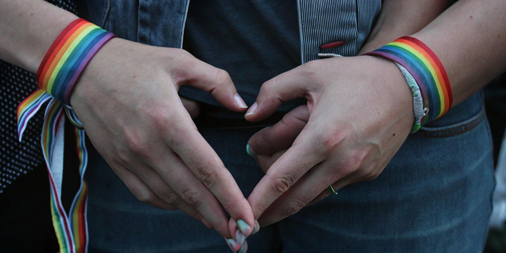 Two hands wearing rainbow-colored bracelets come together to form a heart shape.