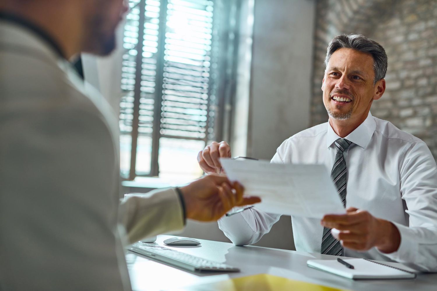 Two people exchange documents across a desk in a bright office; one person smiles while receiving a paper.