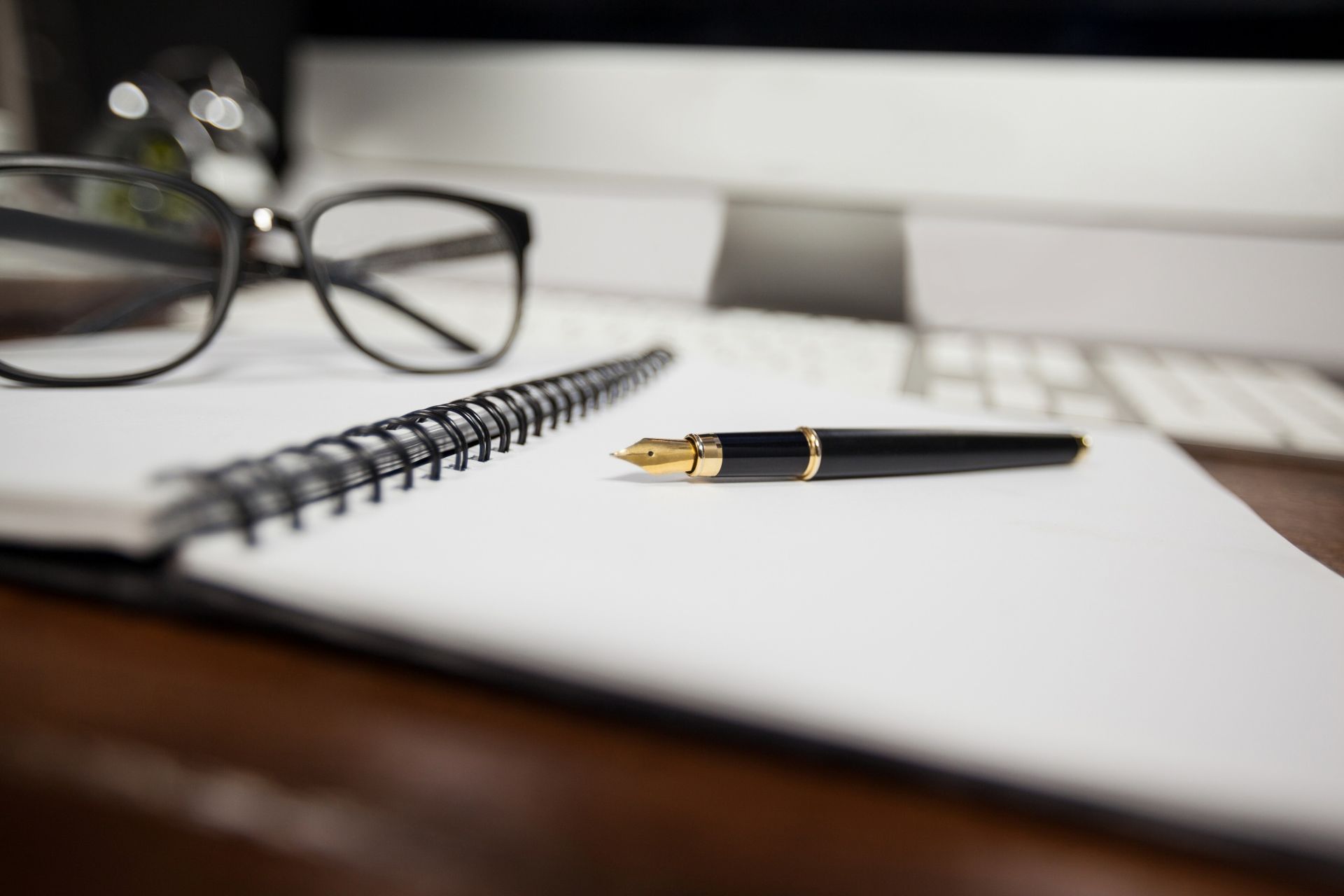 A fountain pen lies on an open spiral notebook next to a pair of black-rimmed glasses on a desk.