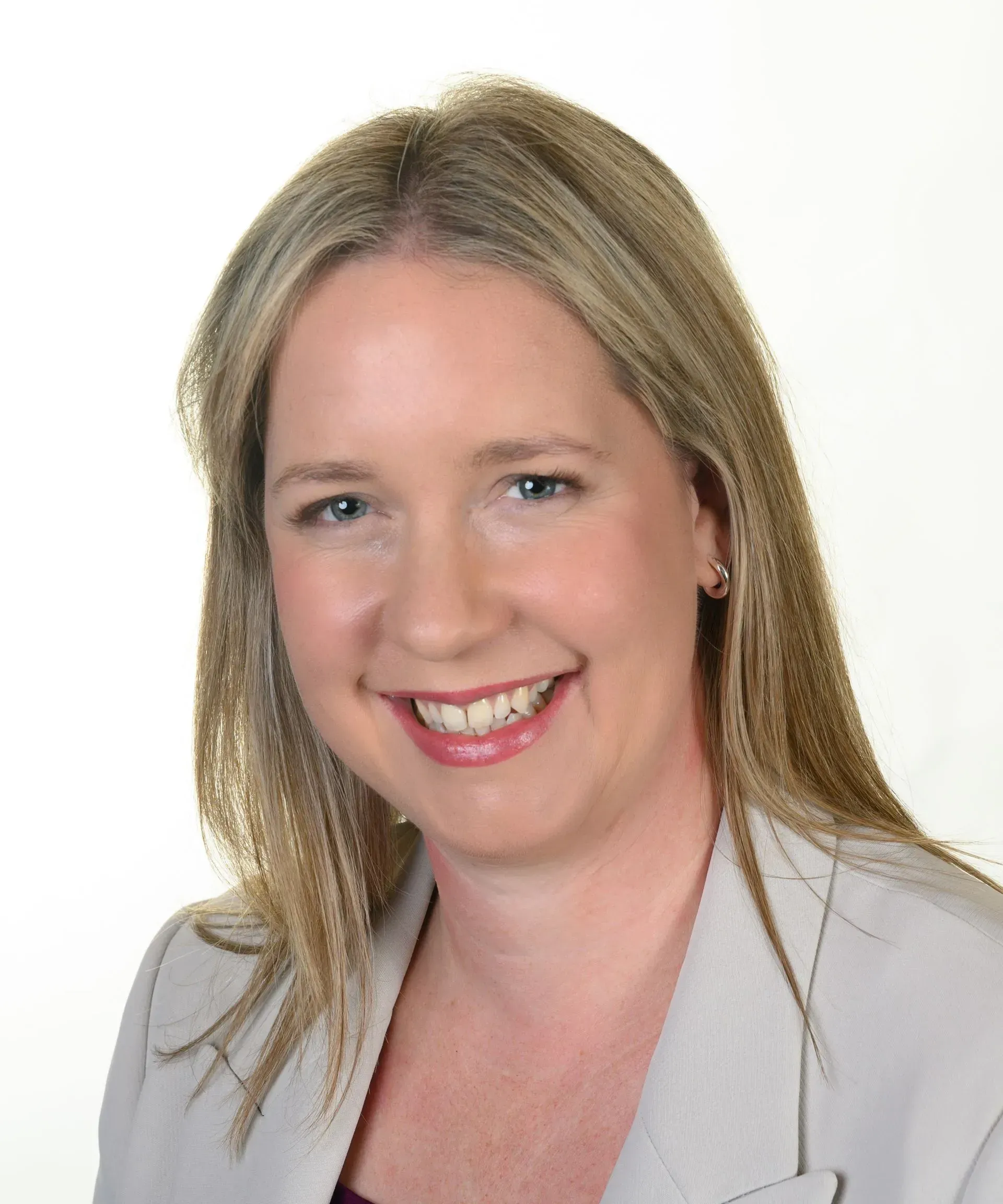 A smiling professional with long blonde hair, wearing a light gray blazer against a plain white background.