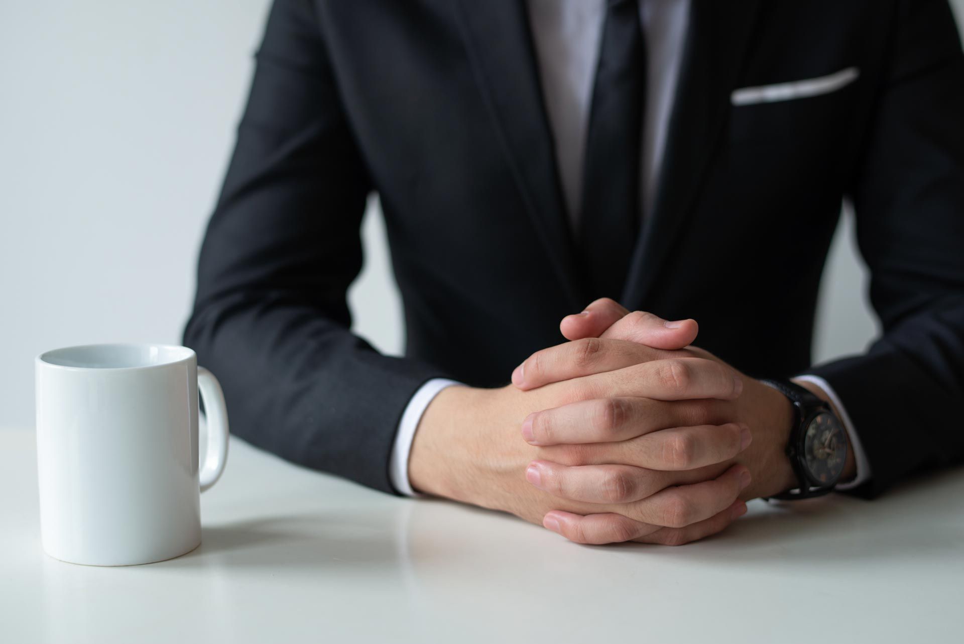 A person in a black suit sits at a white desk with their hands clasped next to a white coffee mug.