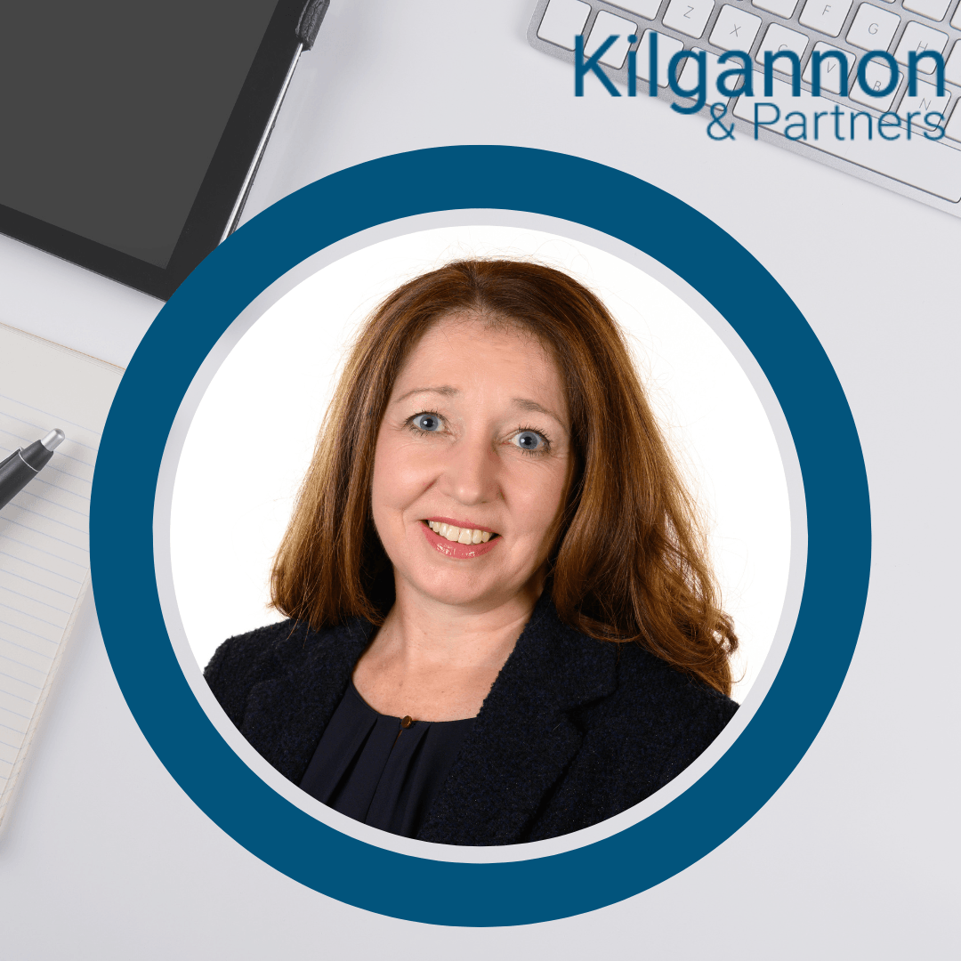 Professional headshot of a smiling person in a black blazer, framed in a circle against a desk background with logo.