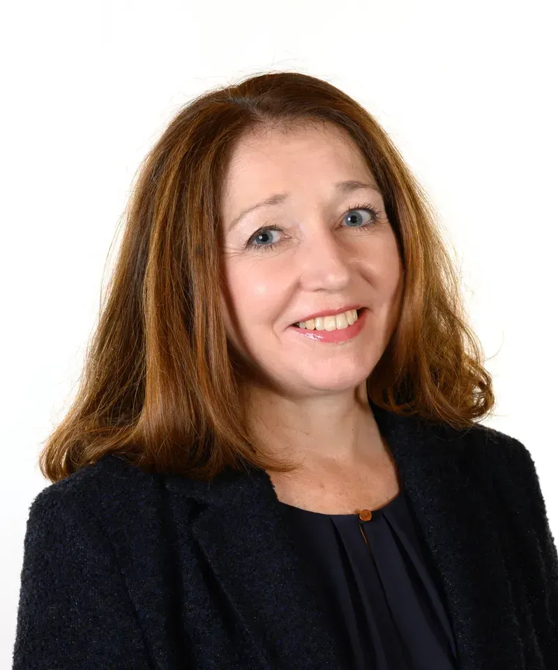 Headshot of a smiling person with shoulder-length brown hair, wearing a dark navy blazer over a dark top.