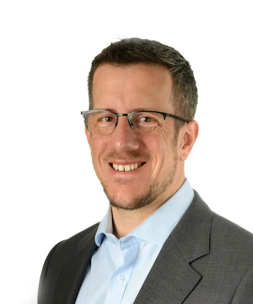 A person with short hair and glasses wearing a light blue shirt and dark suit jacket, smiling against a white background.