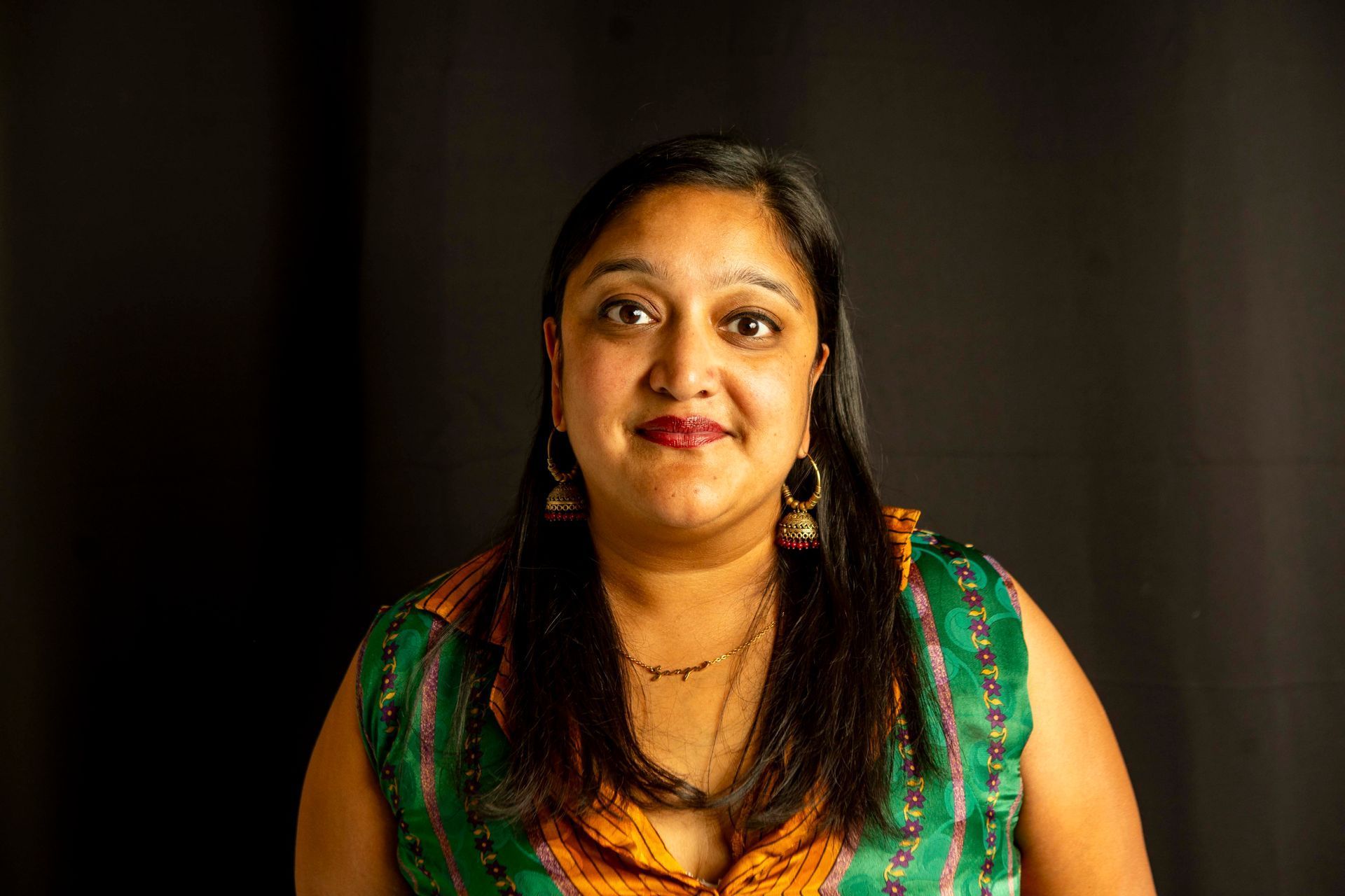 A person with long dark hair, wearing a green and orange patterned top, looks toward the camera against a black background.