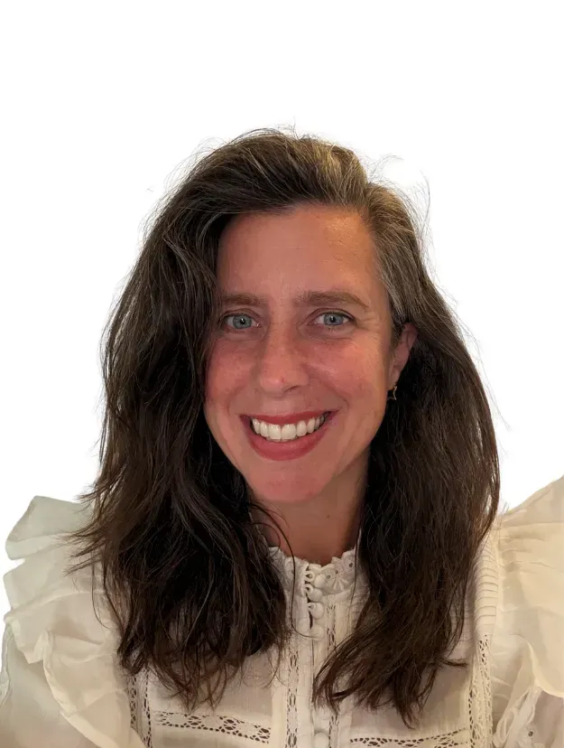 A person with shoulder-length, dark wavy hair, smiling at the camera, wearing a white blouse with ruffled sleeves.