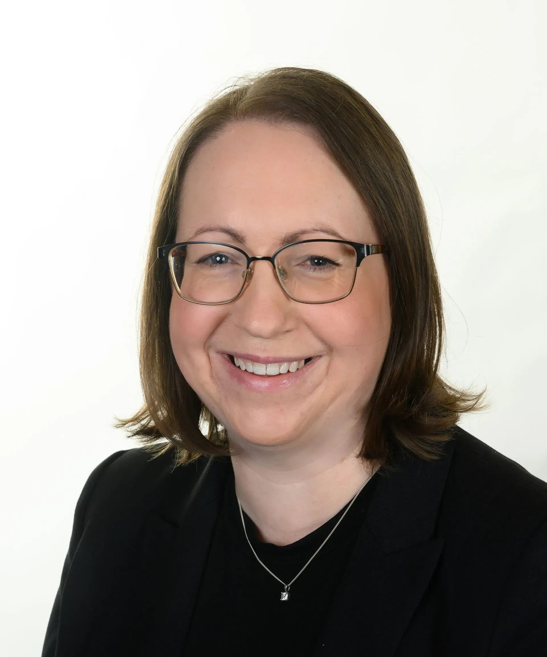 A person with shoulder-length brown hair wearing glasses and a black blazer, smiling against a plain white background.