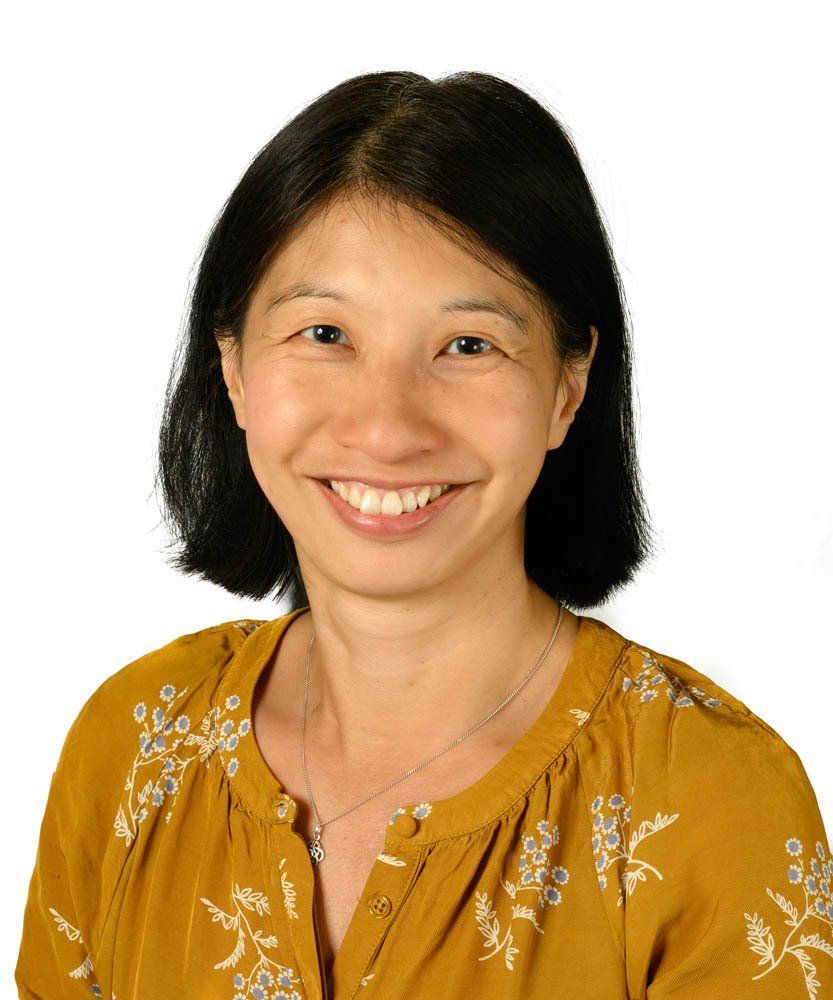 A person with dark hair smiling at the camera, wearing a mustard yellow floral blouse and a silver necklace.