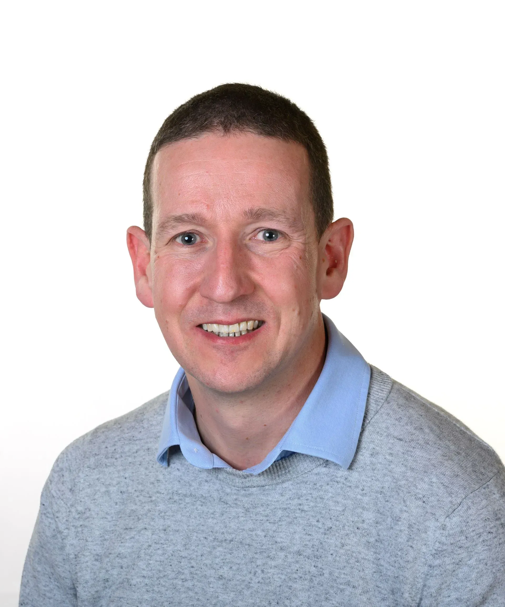 A person with short, dark hair, wearing a light blue collared shirt under a gray sweater, smiling against a white background.