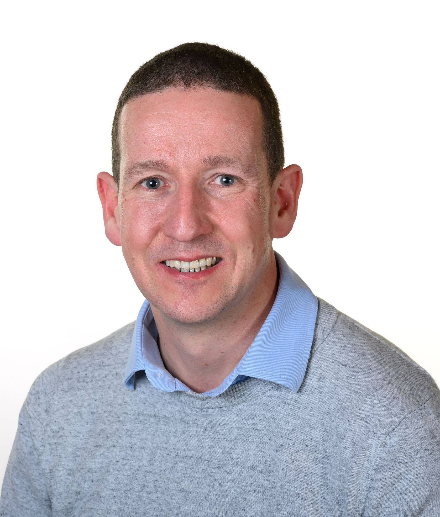 A smiling individual wearing a light blue collared shirt under a gray sweater against a white background.