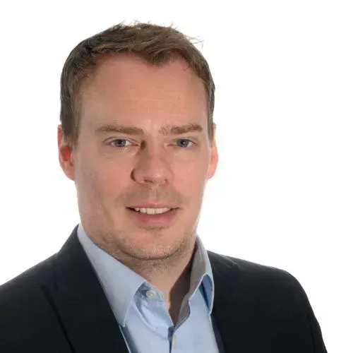 A person with short hair, wearing a dark blazer over a light blue button-down shirt, smiles against a white background.