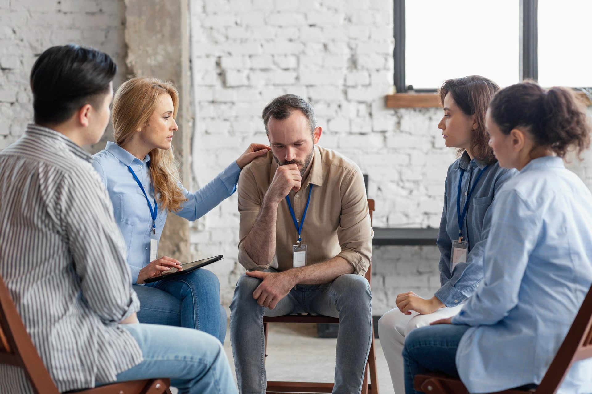 A group sits in a circle for a support session; one person rests a hand on a distressed individual's shoulder.
