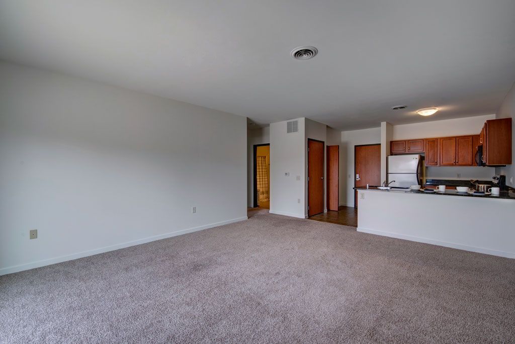 An empty living room with a carpeted floor and a kitchen in the background